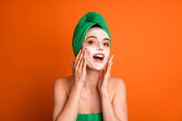 Photo of sweet shiny lady wear green towel head applying soap two arms cheekbones isolated orange color background - Foto, Imagen