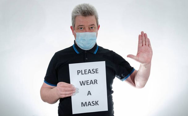 Egy orvosi maszkot viselő férfi táblát tart a mondattal, kérem, viseljen maszkot, miközben tenyérrel nyújtva fogja a kezét, hogy megálljon. - Fotó, kép