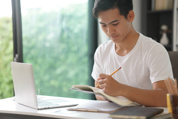 азиатский молодой студент-подросток предприниматель-подросток, работающий с компьютером, пишет записку, изучая обучение онлайн дома. Вебинар по электронному обучению. социальная дистанция в ковидной пандемии - Фото, изображение
