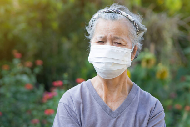 Egy idős nő, aki sebészeti maszkot visel, miközben a kertben áll. Maszk a vírus, a koronavírus, a kovid-19, a baktériumok, a pollen magvak és még sok más védelmére. Az idősek és az egészségügyi ellátás fogalma. - Fotó, kép