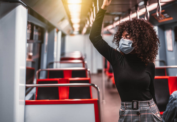 Portrait of a curly young black female in a virus protective mask and a turtleneck, holding onto the handrail of a subway train carriage with a row of empty red seats and a copy space area on the left - Valokuva, kuva