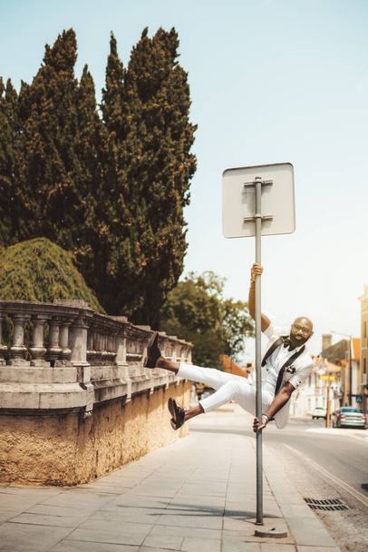 A fancy bald bearded black guy in glasses and an elegant white suit is hanging on a road sign; a fashionable African man entrepreneur is fooling around and jumped up and grabbed the pole near the road - Фото, зображення