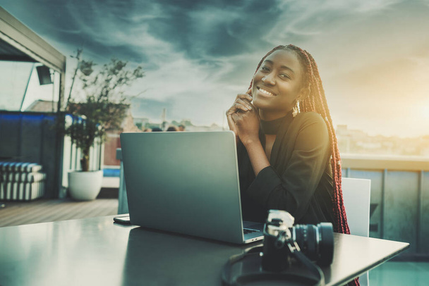 A portrait of a ravishing cheerful young black female freelancer smiling and enjoying an evening while sitting at the table of a street cafe on the roof with a laptop and film camera in front of her - Photo, image