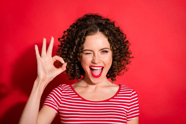 Photo of adorable stunning curly young lady wear casual striped outfit blinking showing okey sign isolated red color background - Fotoğraf, Görsel
