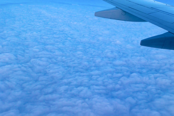 Blick auf den blauen bewölkten Himmel aus der Höhe des Fluges über den Wolken aus dem Flugzeugfenster. Teil eines Flugzeugflügels im Rahmen - Foto, Bild