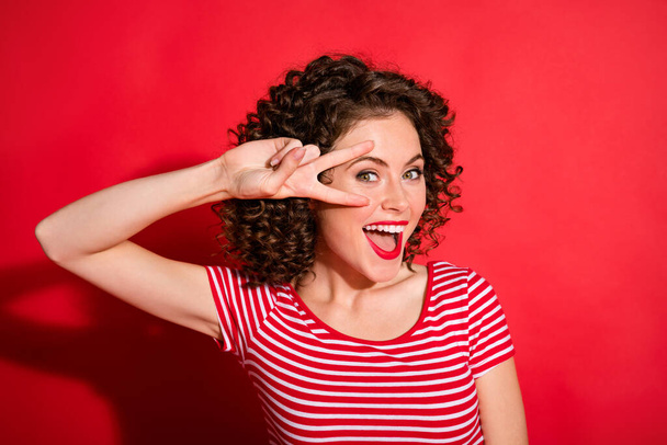 Photo of sweet funny wavy young woman dressed striped outfit showing v-sign eye isolated red color background - Foto, Bild