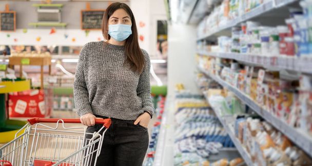 Dívka v ochranné masce stojí s nákupním vozíkem v blízkosti ledniček s mléčnými výrobky v supermarketu. Místo pro text. - Fotografie, Obrázek