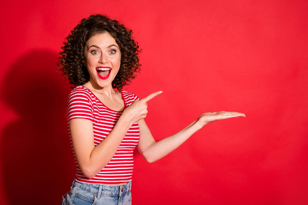 Photo of surprised wavy young woman dressed striped outfit pointing empty space demonstrating product isolated red color background - Foto, Bild