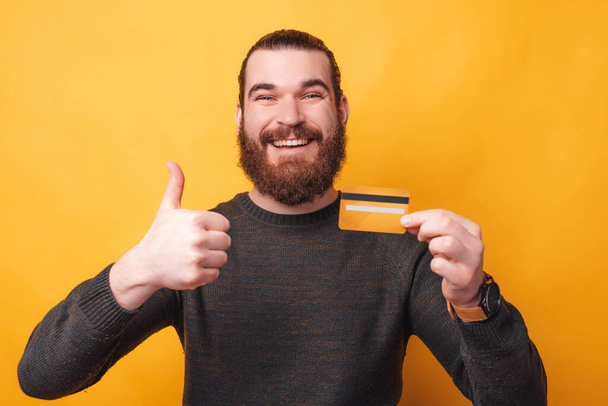 Retrato del joven sonriente sosteniendo tarjeta de crédito y mostrando el pulgar hacia arriba - Foto, imagen