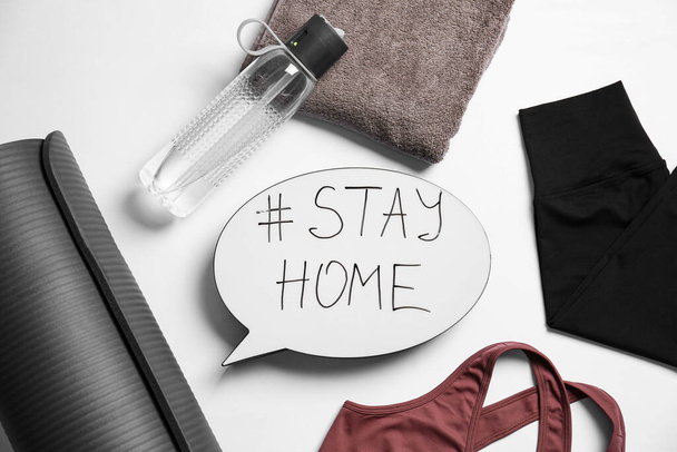 Stylish sportswear, yoga mat and speech bubble with hashtag Stay at Home on white background, flat lay. Self isolation during ccovid19 pandemic - Photo, image