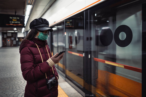 街中で通勤やホームで電車を待っている間にスマートフォンを使用して保護顔マスクを持つ若いアジアの女性 - 写真・画像