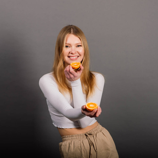 Smiling woman eating orange in studio, half fruit, blonde long hair. - Photo, Image