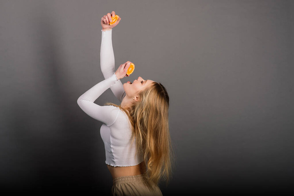 Mujer sonriente comiendo naranja en el estudio, media fruta, pelo largo rubio. - Foto, imagen