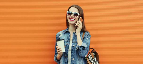 Retrato de una joven sonriente llamando a un teléfono inteligente con una chaqueta de mezclilla sobre un fondo naranja - Foto, Imagen