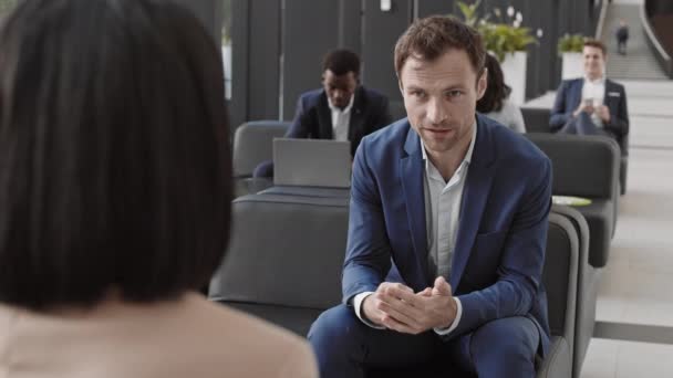 Achteraanzicht van knappe blanke zakenman in elegant blauw pak zittend op de bank voor onherkenbare vrouwelijke collega, in gesprek en vervolgens kijkend naar de camera - Video