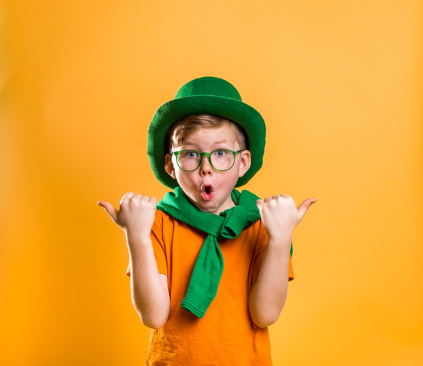 聖パトリックの日。緑の帽子、眼鏡、黄色で隔離されたセーターでスタイリッシュな小さな子供の少年の肖像画。緑のレプション。驚いた子供の顔を指を指している。販売。割引よ。3月. - 写真・画像