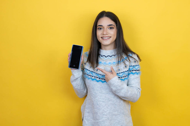 Young caucasian woman wearing casual sweater over yellow background holding smartphone showing screen smiling happy pointing with hand and finger - Photo, Image