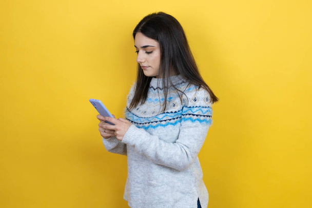 若いですcaucasian女性身に着けていますカジュアルセーター上の黄色背景テキスト上の彼女の携帯電話 - 写真・画像