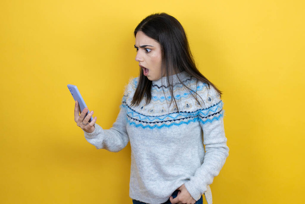 Молодая белая женщина в случайном свитере на жёлтом фоне разговаривает по телефону с беспокойным выражением лица - Фото, изображение