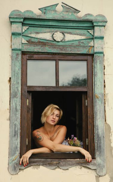 Art portrait of a beautiful young woman, looks through an old window peeps out, sits on the windowsill in grunge style. - Фото, зображення