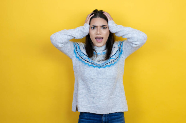 Young caucasian woman wearing casual sweater over yellow background crazy and scared with hands on head, afraid and surprised of shock - Foto, Bild