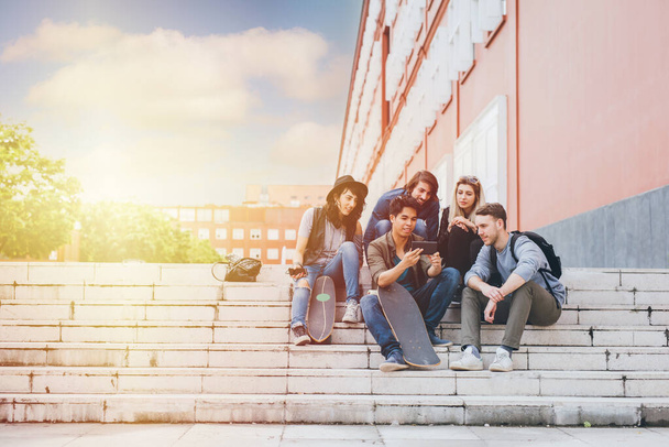 Grupo de amigos multiétnicos sentados al aire libre luz de fondo millennials usando teléfonos inteligentes adictos a las tendencias tecnológicas siguiendo y charlando y viendo videos - Foto, imagen