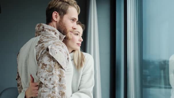 Ελκυστικό ζευγάρι νεαρών Αμερικανών στρατιωτών άντρας και ξανθιά γυναίκα αγκαλιάζονται ενώ κοιτάζουν το παράθυρο που στέκεται μέσα στα διαμερίσματα - Πλάνα, βίντεο