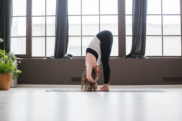 Молодая женщина с длинными косичками в спортивной одежде практикует йогу в яркой комнате с большими окнами. Красивая девушка практикует асану Падангушт йогу. - Фото, изображение