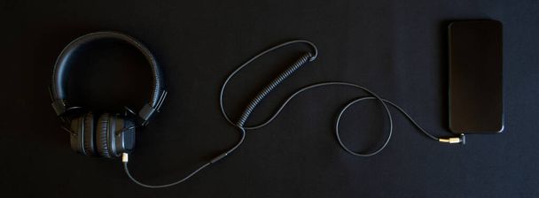 Musta langallinen kuulokkeet korva tyynyt ja älypuhelin makaa mustalla taustalla. Analoginen johto yhteys mini-jack 3,5 mm. Musiikkia ja luovuutta. Banneri - Valokuva, kuva