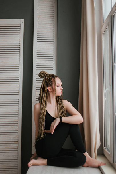 Young woman with long braids in sportswear practices yoga. A beautiful girl is sitting near a large window. Large studio hall - Fotó, kép