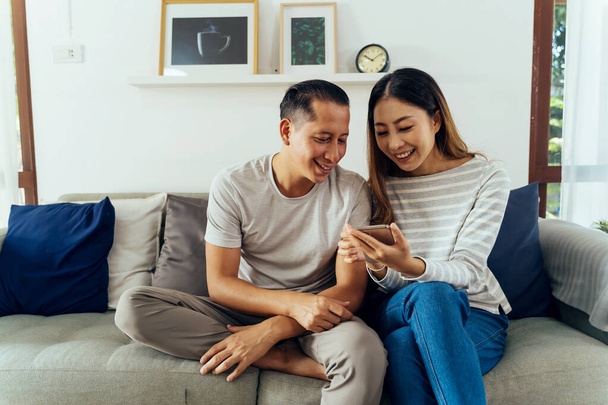 30s young adult Asian man and woman playing a mobile phone together sits on sofa in cozy living room at home. Happy couple internet user in casual clothes on couch. Technology usage in Asia concept - Фото, зображення