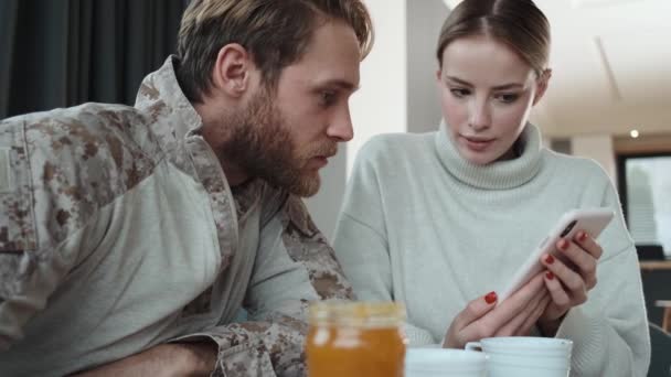 Bella coppia giovane soldato americano uomo e donna bionda stanno utilizzando smartphone seduto all'interno degli appartamenti - Filmati, video