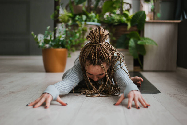Jonge vrouw met lange vlechten in sportkleding die yoga beoefent in een lichte kamer met grote ramen. Mooi meisje oefent asana hatha yoga. Grote hal met planten - Foto, afbeelding