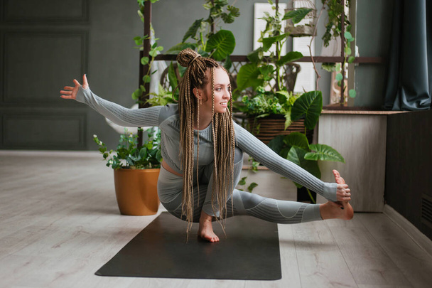 Mujer joven con trenzas largas en ropa deportiva practicando yoga en una habitación luminosa con grandes ventanales. Hermosa chica practica asana hatha yoga. Gran salón con plantas - Foto, Imagen