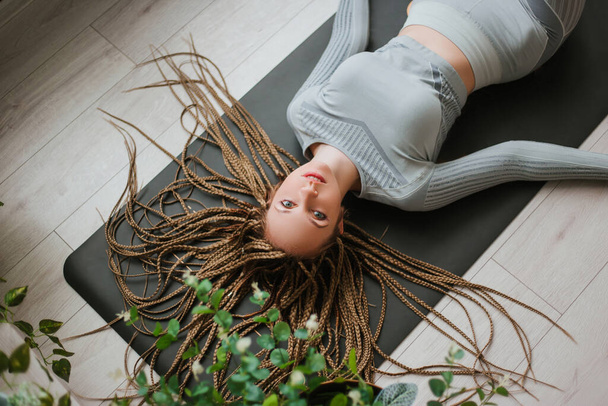 Young woman with long braids in sportswear practices yoga. A beautiful girl lies and rests after yoga. Big hall - Photo, Image