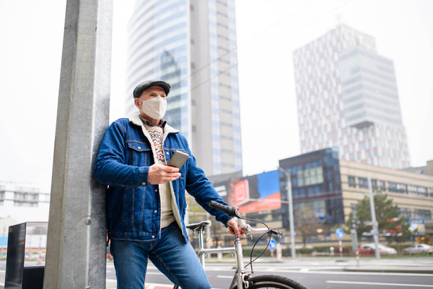 Senior man with bicycle and smartphone outdoors on street in city, coronavirus concept. - Fotografie, Obrázek