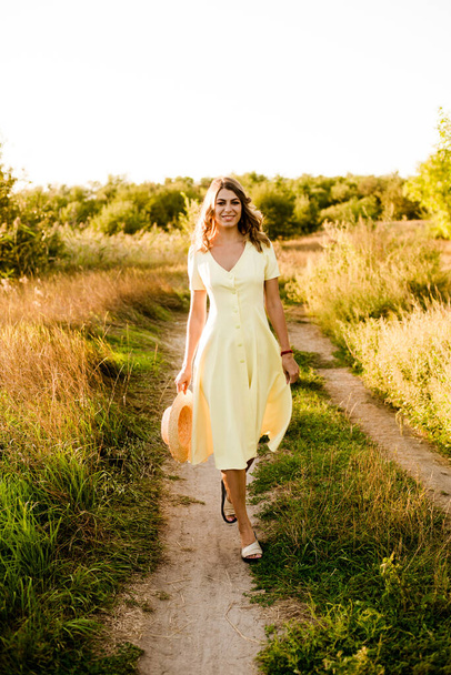 A young beautiful girl with blond curly hair in a light summer yellow dress and with a straw hat in her hands in a field in the countryside at sunset. People and nature  - Foto, imagen