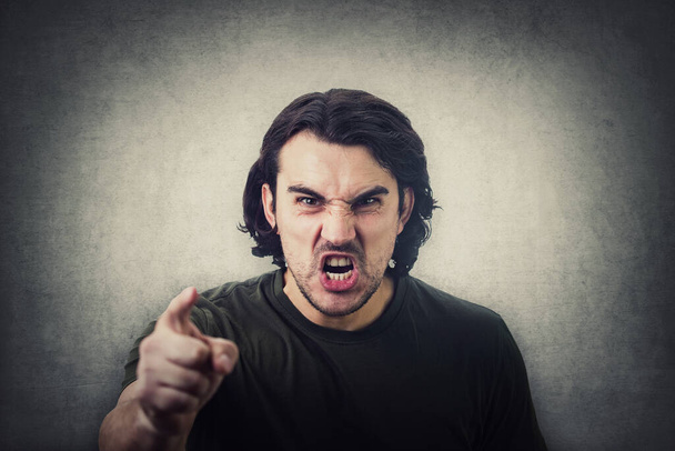 Furious young man pointing forefinger to camera blaming someone as guilty, or scolding isolated on grey wall background. Angry guy screaming and showing with index being annoyed - Photo, Image