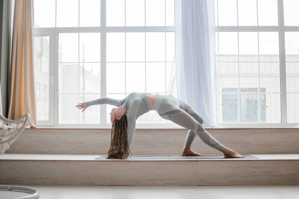 Spor kıyafetleri içinde uzun örgülü genç bir kadın geniş pencereli parlak bir odada yoga yapıyor. Güzel bir kız hatha yogası yapıyor.. - Fotoğraf, Görsel