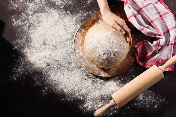 Homemade wholemeal dough for bread and female hand on wooden table with flour, rolling-pin, dark rustic background. Top view, copy space. Alternative handmade food cooking concept. - Photo, Image