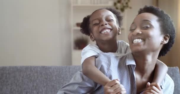 Αφρικανική αμερικανική νεαρή γυναίκα μαμά μεγαλύτερη αδελφή νταντά κάθεται στον καναπέ στο σπίτι αγκαλιάζει λίγο χαριτωμένο κορίτσι προσχολικής ηλικίας αστείο ποζάρουν μπροστά από κάμερα μαζί χαμογελαστά δόντια χαχανίζουν ευρέως, το πρόσωπο από κοντά - Πλάνα, βίντεο