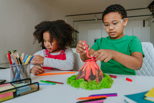 Сестра и брат с удовольствием делают из детей модель DIY volcano, играя в проект Clay for school. Концепция домашнего образования. - Фото, изображение