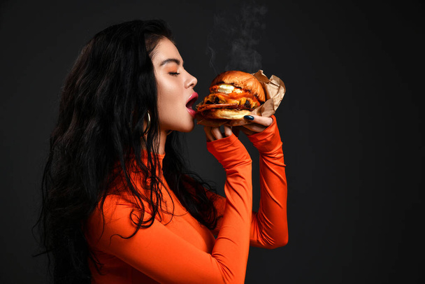 Portret van hete sexy brunette vrouw met pruillip in rode kleren eten bijten grote smakelijke ambachtelijke hamburger met stoom - Foto, afbeelding