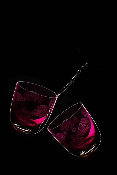 Zwei lila Gläser mit Fischzeichnungen. Schräges Glas unten auf schwarzem Hintergrund. Der links mit einem Spritzer Flüssigkeit und der rechts leer. Kopierraum - Foto, Bild