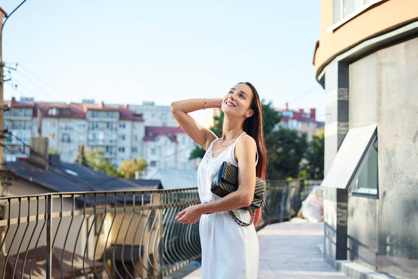 Young brunette woman, wearing silk white dress, holding black purse,standing on hotel balcony in the morning. Female portrait in summer. Fashion model posing on terrace with beautiful city view. - Foto, afbeelding