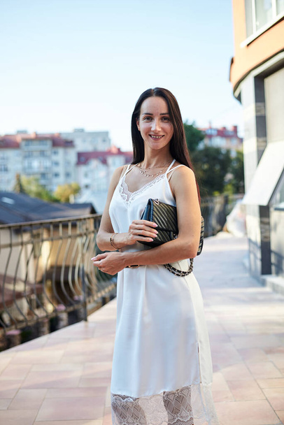Young brunette woman, wearing silk white dress, holding black purse,standing on hotel balcony in the morning. Female portrait in summer. Fashion model posing on terrace with beautiful city view. - Photo, image