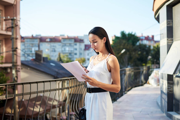Young brunette business woman, wearing silk white dress, holding sheets of paper,standing on hotel balcony. Female portrait in summer. Fashion model posing on terrace with beautiful city view. - Fotoğraf, Görsel