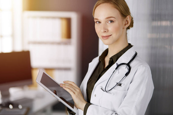 Cheerful smiling female doctor using tablet computer in sunny clinic. Portrait of friendly physician woman at work. Perfect medical service in hospital. Medicine concept - Photo, image