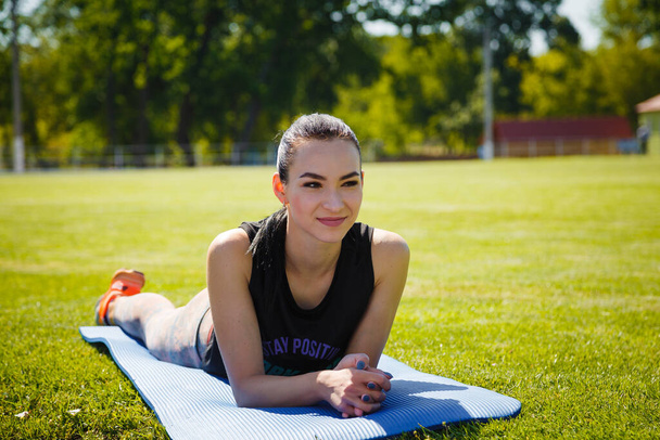 Young slim girl in sport clothes lying on yoga mat on stadium green lawn. Sport, healthy way of life, fitness concept. - Photo, Image