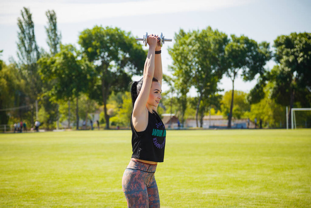 Улыбающаяся молодая женщина, стоящая на зеленой лужайке стадиона с гантелями в руках до небес. Спорт, физкультура, здоровый образ жизни, концепция фитнеса. - Фото, изображение